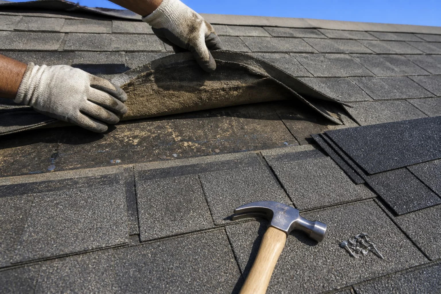 Gloved hands lifting an asphalt shingle with a hammer nearby